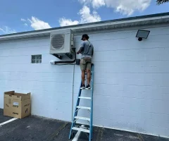 A man on a ladder servicing a wall-mounted air conditioner unit on a white building exterior in a parking lot