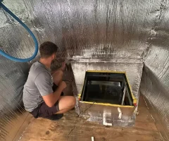 A man installing reflective foil attic insulation around a skylight opening on a plywood attic floor.