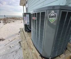 Outdoor HVAC units on a raised wooden platform beside a beach house, sand dunes and ocean in background