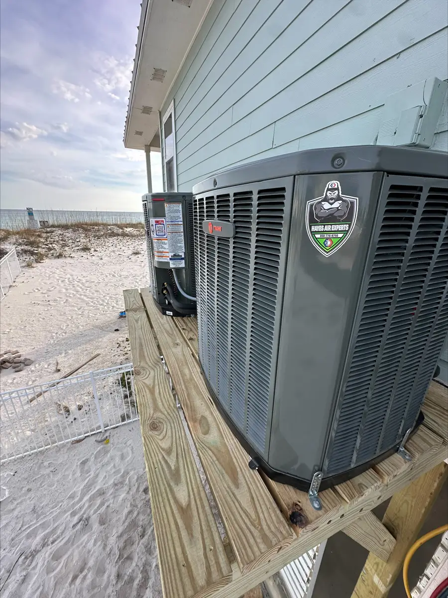 Outdoor HVAC units on a raised wooden platform beside a beach house, sand dunes and ocean in background