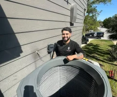 A man HVAC technician smiling while servicing an outdoor air conditioning unit beside a house siding