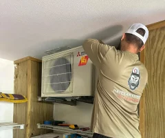 A man installing a Mitsubishi Electric mini-split HVAC unit on a wall inside a wooden utility room.