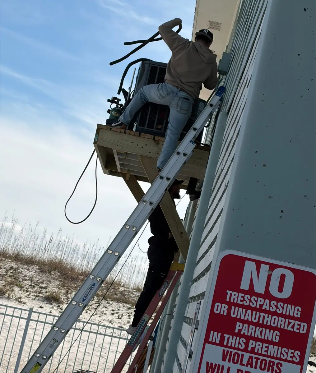 Worker on ladder installing HVAC unit on exterior wall from a wooden platform by a sandy beach.
