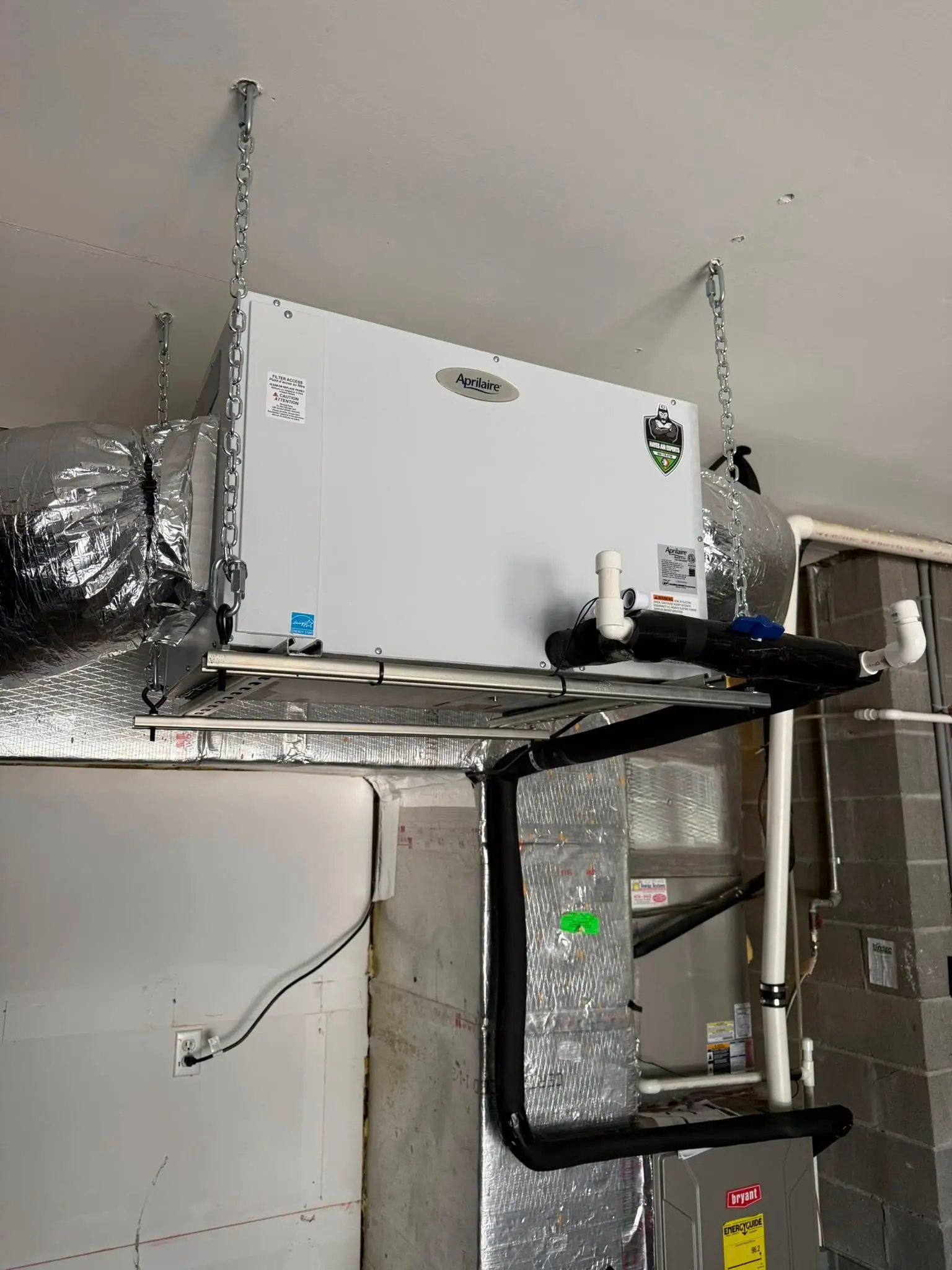 Aprilaire HVAC unit suspended from basement ceiling by chains, with insulated ductwork and condensate drain.