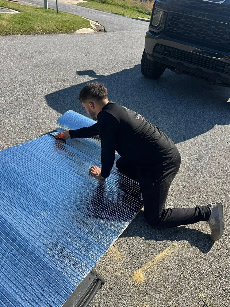 Man kneeling on pavement cutting reflective foil insulation mat beside a black pickup truck