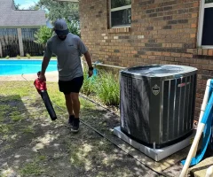 A man using a leaf blower to clear debris near an outdoor air conditioner beside a brick house and backyard pool