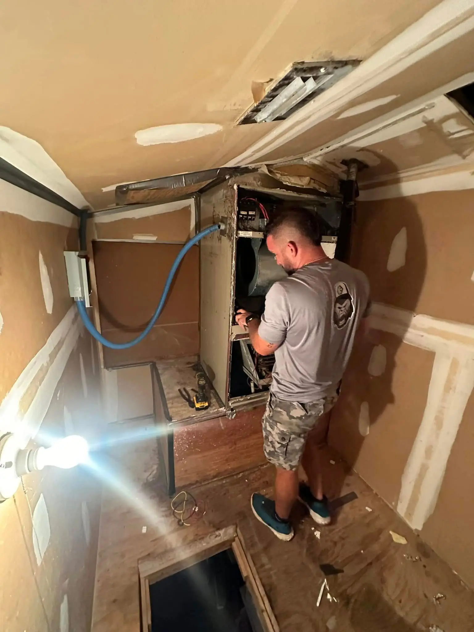 A man repairing an HVAC unit in a narrow unfinished room with exposed drywall, tools and a blue flexible duct
