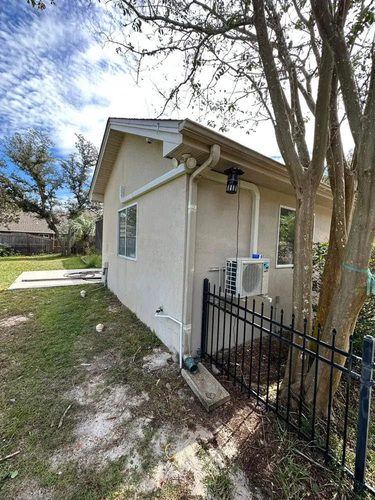 Side view of beige stucco house with wall-mounted mini-split AC, gutters, black metal fence and tree in a grassy side yard.