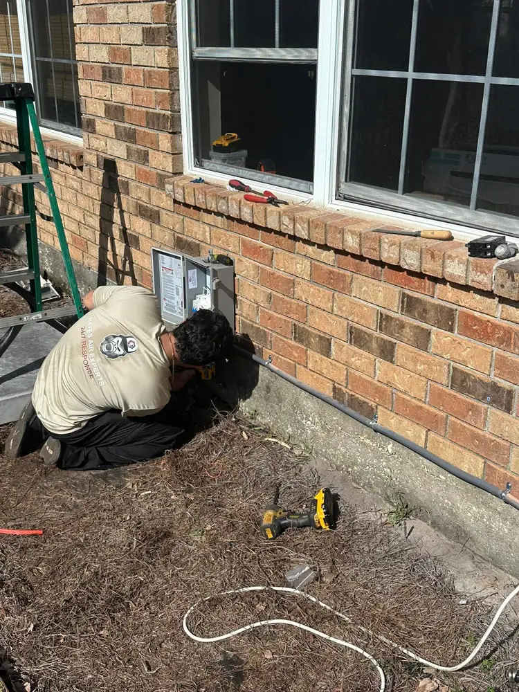 A man kneeling beside a brick house repairing an outdoor electrical box with drill, tools, and ladder nearby