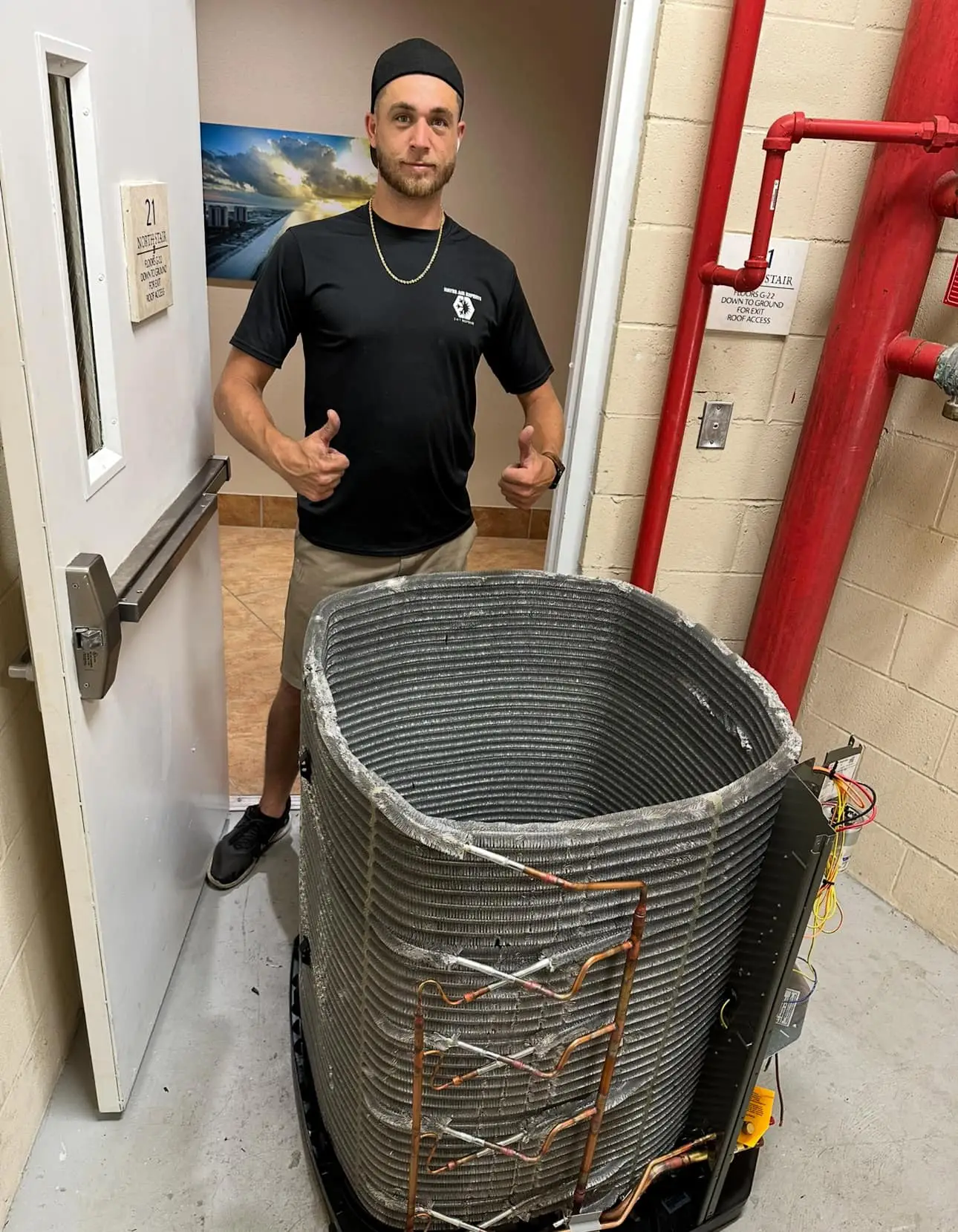 A man giving thumbs up next to a removed HVAC condenser coil in a utility room with red pipes