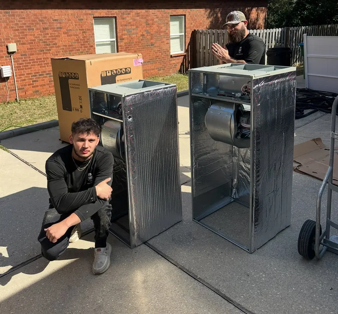 Two men with HVAC blower housings on a driveway, one man crouching beside units in front of a brick house