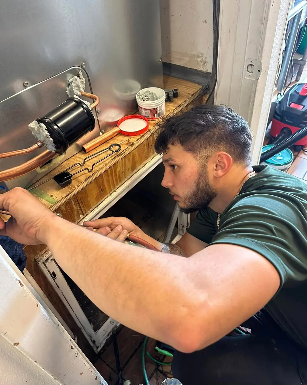 Man kneeling and repairing refrigerator compressor and copper tubing inside appliance cabinet.