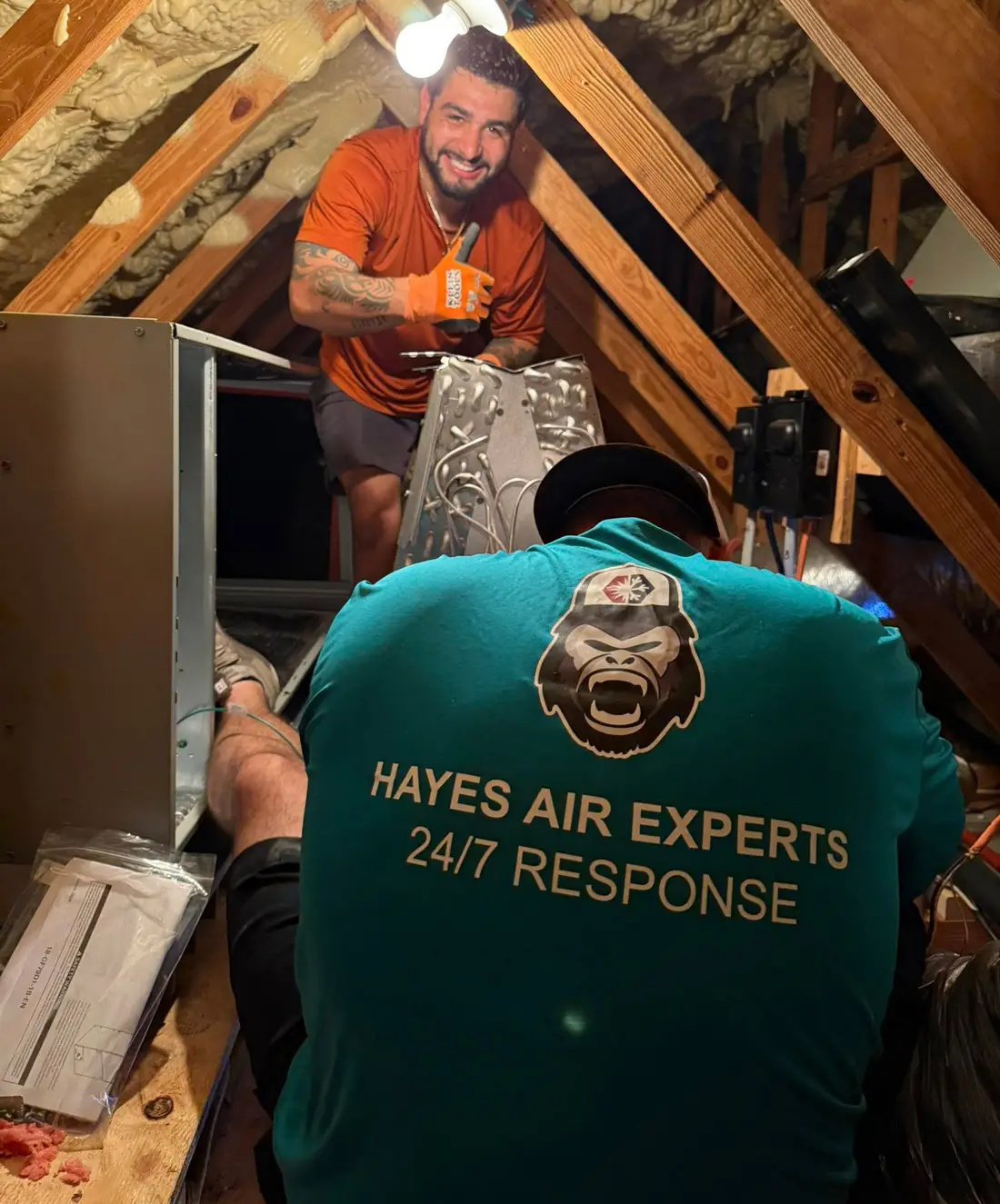 Two HVAC technicians in an attic; one kneels wearing 'Hayes Air Experts' shirt, other flashes thumbs up